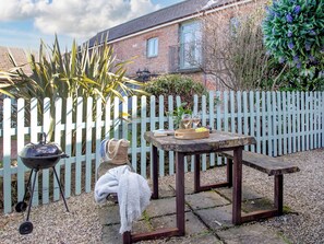 Un jardí amb una barbacoa, una taula amb una cistella de fruita i una banca.
