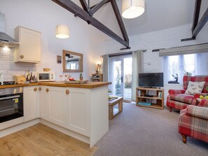 A modern kitchen with white cabinets, a wooden countertop, and a built-in oven. There is a dining area with a red plaid sofa and a television. - Rose Cottage - UKC6181 (Marldon, near Paignton)