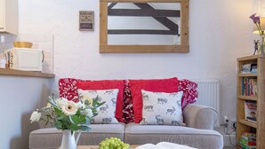 Ein gemütliches Wohnzimmer mit einer Couch, einem Couchtisch mit einem Buch und Trauben, einer Vase mit Blumen und einem Bücherregal.