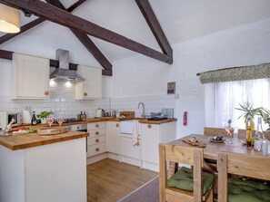 A kitchen with wooden beams, white cabinets, a dining table with chairs, and a window with curtains. - Mill Cottage - UKC6180 (Marldon, near Paignton)
