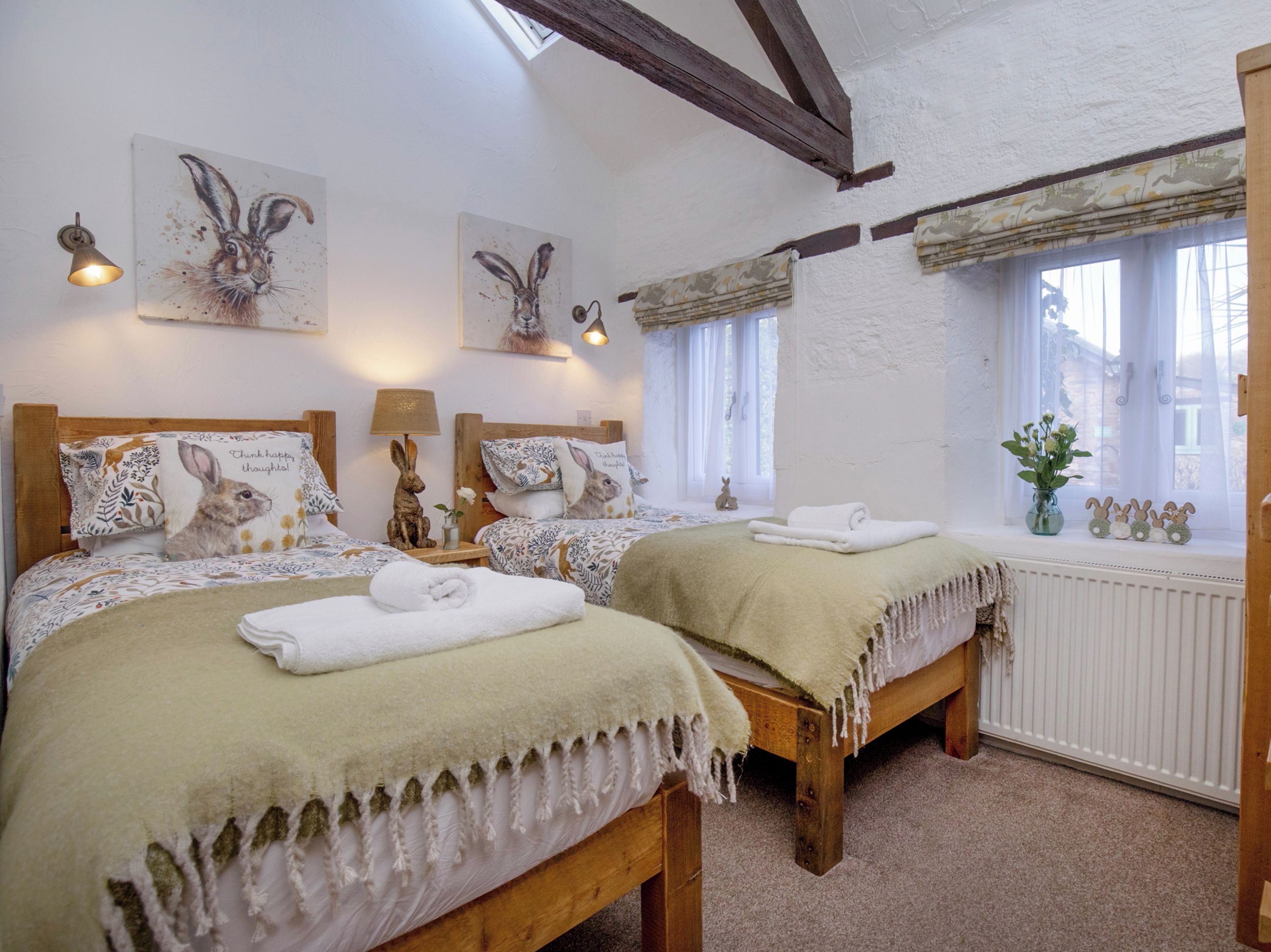 Une chambre avec deux lits en bois, chacun agrémenté d’un oreiller orné d’un motif représentant un lapin, une petite table avec une lampe et une fenêtre donnant sur l’extérieur.