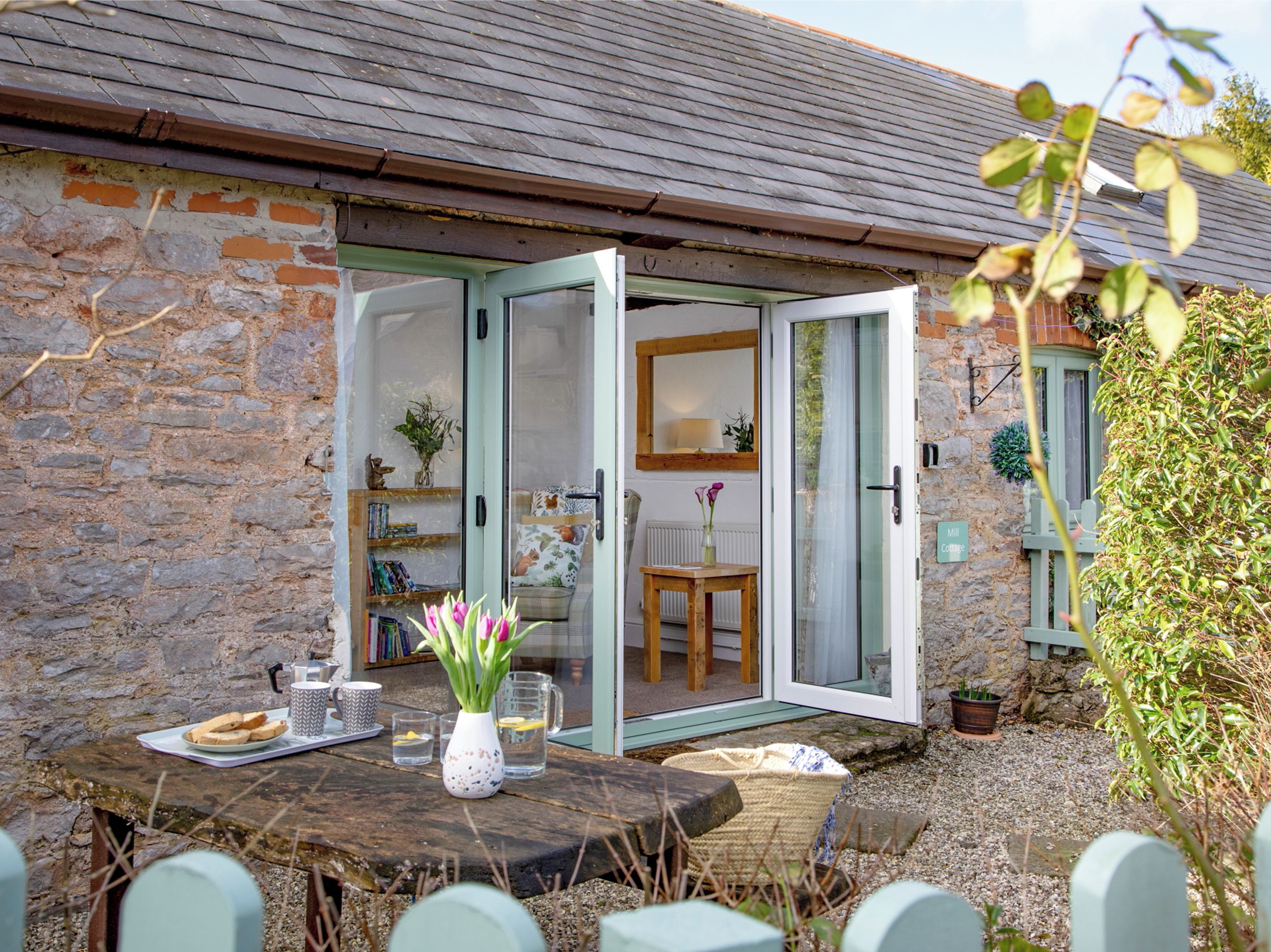 A stone building with a wooden table set for breakfast, a glass door leading inside, and a small garden area.