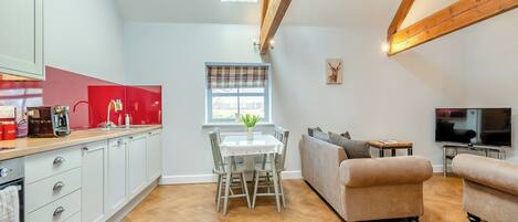 A modern kitchen with white cabinets and a red countertop, a dining area with a white table and chairs, a beige sofa, and a TV in a living room with wooden beams on the ceiling.