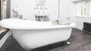 Een klassiek ligbad op poten in een moderne badkamer, met een witte wastafel, een ronde spiegel en een raam met jaloezieën.