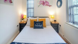 Un dormitorio con una cama, dos mesitas de noche, una ventana con persianas y adornos en la pared, incluidas orquídeas y un espejo circular.