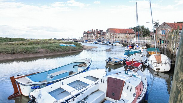 A harbor with boats docked along the water's edge, a building with a red roof in the background, and a grassy area on the left. - A cute and characterful cottage in a charming coastal port. (Blakeney)