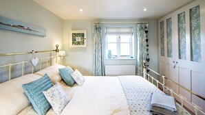 A bedroom with a bed, a window with curtains, a radiator, and a wall with framed pictures. - A cute and characterful cottage in a charming coastal port. (Blakeney)