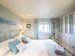 A bedroom with a bed, a window with curtains, a radiator, and a wall with framed pictures. - A cute and characterful cottage in a charming coastal port. (Blakeney)