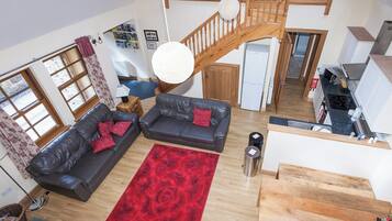 A spacious living area with a wooden staircase, two leather sofas with red cushions, a red floral rug, and a kitchen with modern appliances.