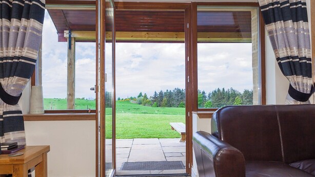 Ein Zimmer mit braunem Ledersofa, einem Holztisch und Blick auf ein grünes Feld durch große Fenster mit gestreifter Vorhang.