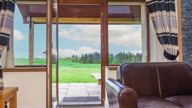 Ein Zimmer mit einer großen Schiebetür aus Glas, die auf eine Terrasse im Freien führt, einem braunen Ledersofa und einem Holztisch.