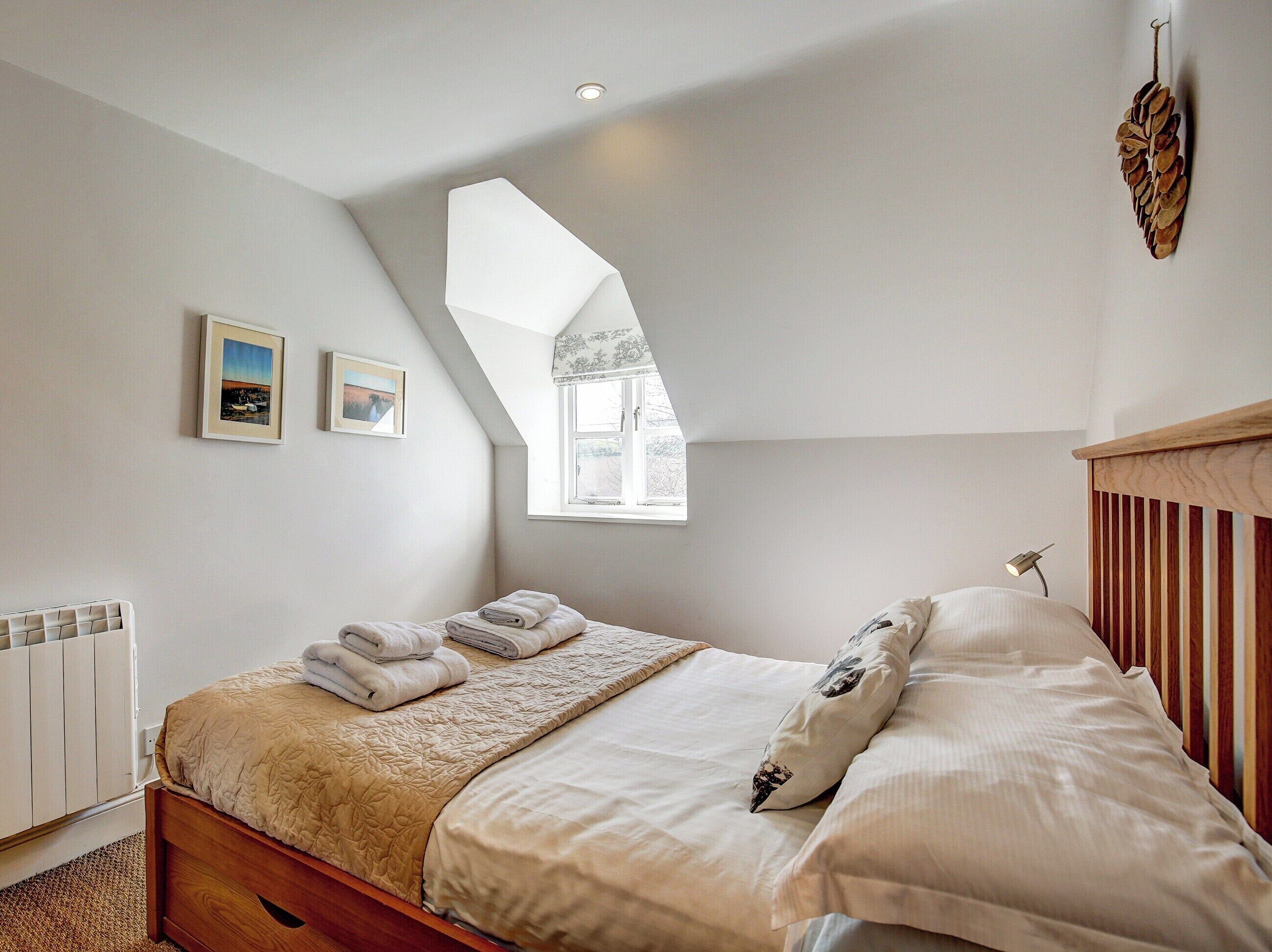 A bedroom with a sloped ceiling, a bed with a wooden headboard, a window, and two framed pictures on the wall.