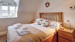 Ein ordentlich bezogenes Bett mit gefalteten Handtüchern, ein hölzernes Kopfteil, ein Nachttisch mit Lampe und ein Fenster mit Blick auf Bäume.