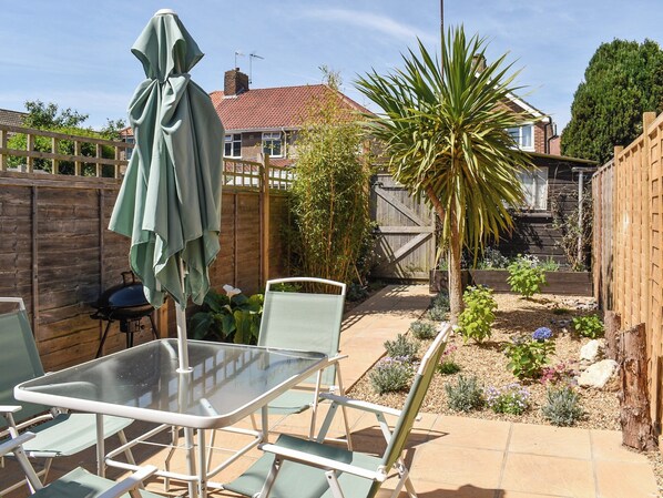 Une terrasse avec une table et des chaises en verre, un parasol, et un jardin agrémenté d’un palmier et de diverses plantes.