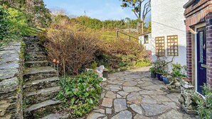 Un sendero de piedra que conduce a un edificio blanco con una puerta azul, rodeado de follaje y un muro de piedra.