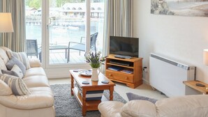 Een woonkamer met een bank, een salontafel, een televisie op een houten meubelstuk en uitzicht op een zwembad en gebouwen door grote ramen.