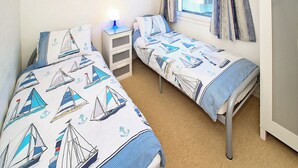 Een kamer met twee enkele bedden, elk voorzien van een bedspreide met zeilbootmotief, een klein nachtkastje met een lamp, en een raam met gordijnen.