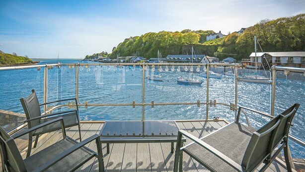 Een terras met een tafel en stoelen met uitzicht op een jachthaven met boten en een heuvel op de achtergrond.