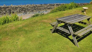 Et picnicbord på en græsplæne med en klippefyldt kystlinje og udsigt til havet og fjerne bjerge.