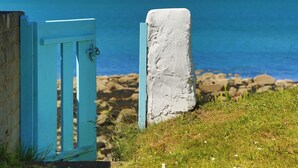 Et kystlandskab med en blå dør og en hvid pæl, der fører til en stenet kystlinje og havet.