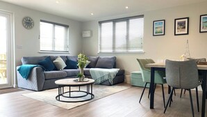 A modern living room with a grey sofa, a round coffee table, and a dining area with a table and chairs. - Holywell - CWC610826 (Perranporth)