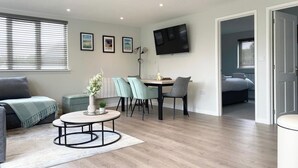 A modern living room with a sofa, a dining table with chairs, a TV mounted on the wall, and a view into the bedroom. - Holywell - CWC610826 (Perranporth)