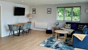 Ein modernes Wohnzimmer mit einem Holzespeisetisch, grauen Stühlen, einem blauen Sofa, einem kleinen runden Tisch und einem an der Wand montierten Fernseher.