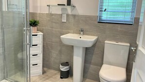 Ein Badezimmer mit Duschkabine aus Glas, einem Standwaschbecken, einer Toilette und einem kleinen Ablagebereich mit einer Pflanze.