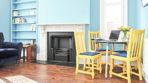 Een woonkamer met een open haard, een boekenkast, een blauwe bank en een eettafel met gele stoelen.