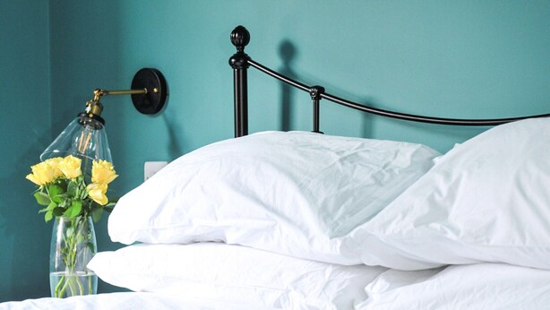 Een bed met witte lakens, een zwart metalen hoofdbord en een glazen vaas met gele bloemen tegen een turquoise muur.