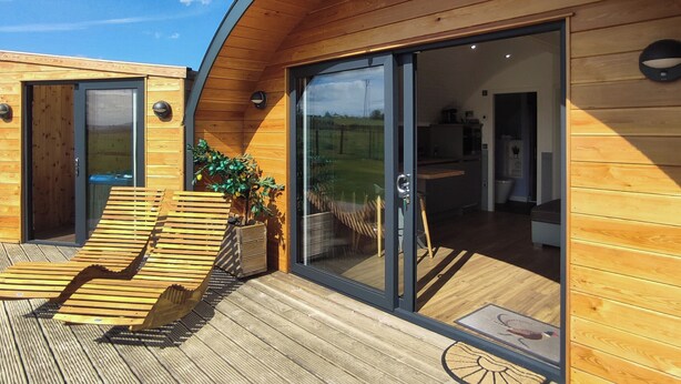 Un'accogliente baita moderna in legno, con una veranda, una porta vetrata che dà su una cucina e un'area relax con una panca e una pianta in vaso.