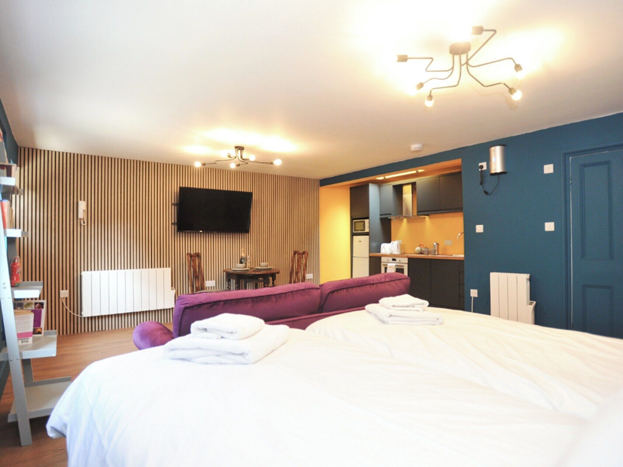 Une chambre d’hôtel moderne dotée d’un grand lit, d’un canapé, d’un téléviseur à écran plat fixé au mur, d’un petit coin cuisine et d’une salle de bain avec un porte-serviettes.