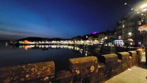 Eine Uferpromenade mit Steinbarriere, ruhigem Wasser, das Lichter spiegelt, und Gebäuden mit beleuchteten Schildern.