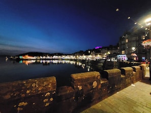 A waterfront promenade with a stone barrier, calm water reflecting lights, and buildings with illuminated signs. - George Street Apartment (Oban)