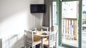 Een eetgedeelte met een tafel voor vier personen, witte stoelen, een flatscreen televisie aan de muur en een schuifdeur naar een buitenruimte.