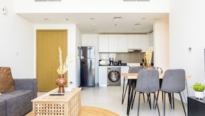 Ein modernes Wohnzimmer mit einem grauen Sofa, einem Holztisch und einem Essbereich mit grauen Stühlen. Im Hintergrund sind eine Waschmaschine und ein Kühlschrank zu sehen.