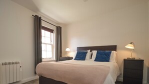 Ein ordentlich eingerichtetes Schlafzimmer mit einem großen Bett, zwei Nachttischen mit Lampen, einem Fenster mit Vorhängen und einem Heizkörper.