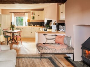A cozy living room with a fireplace, a dining area with wooden furniture, and a kitchen with white cabinets and a window. - Boundary Cottage - UKC6387 (Holbeton)