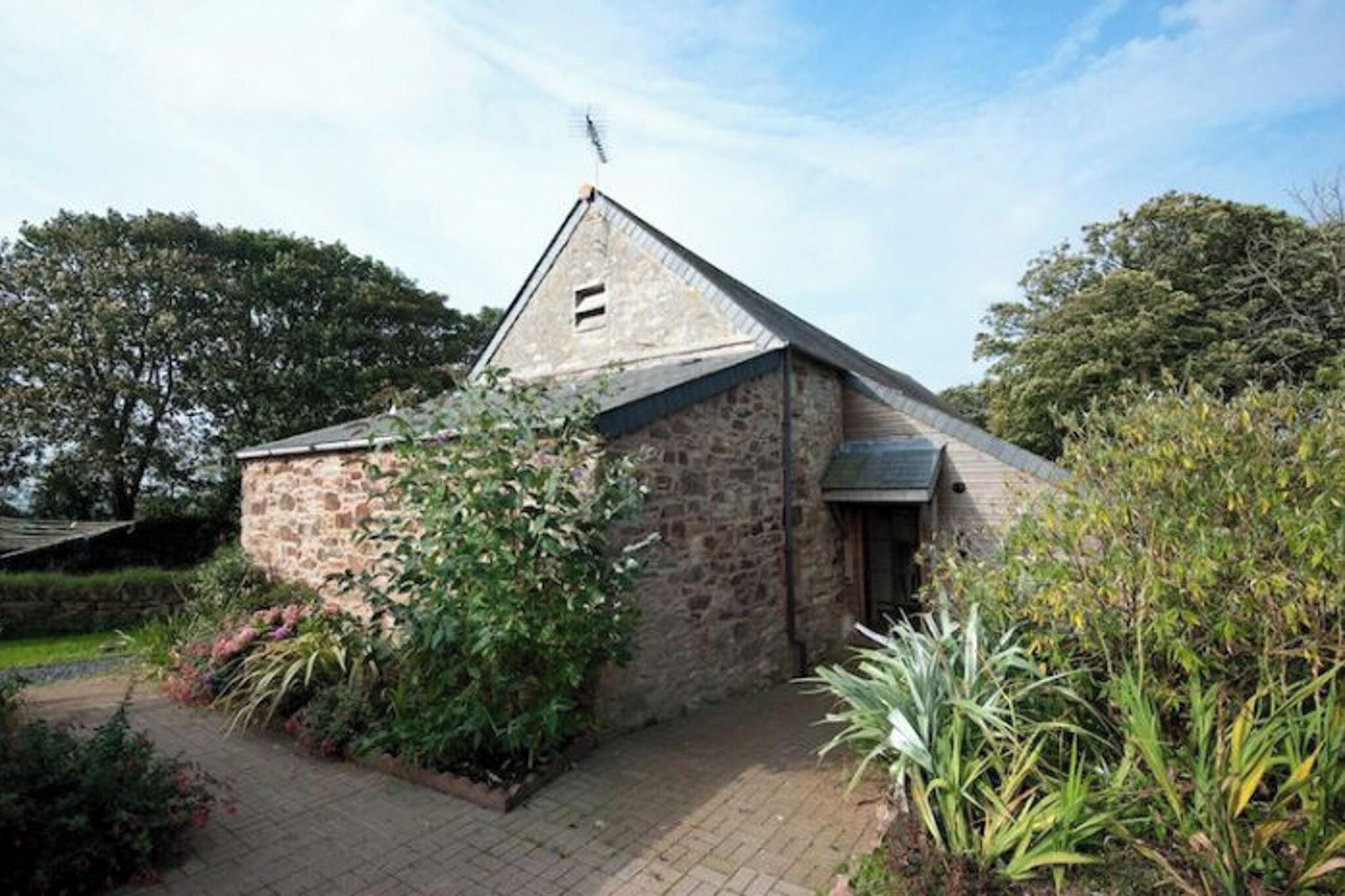 A stone building with a sloped roof, surrounded by greenery and a paved pathway.