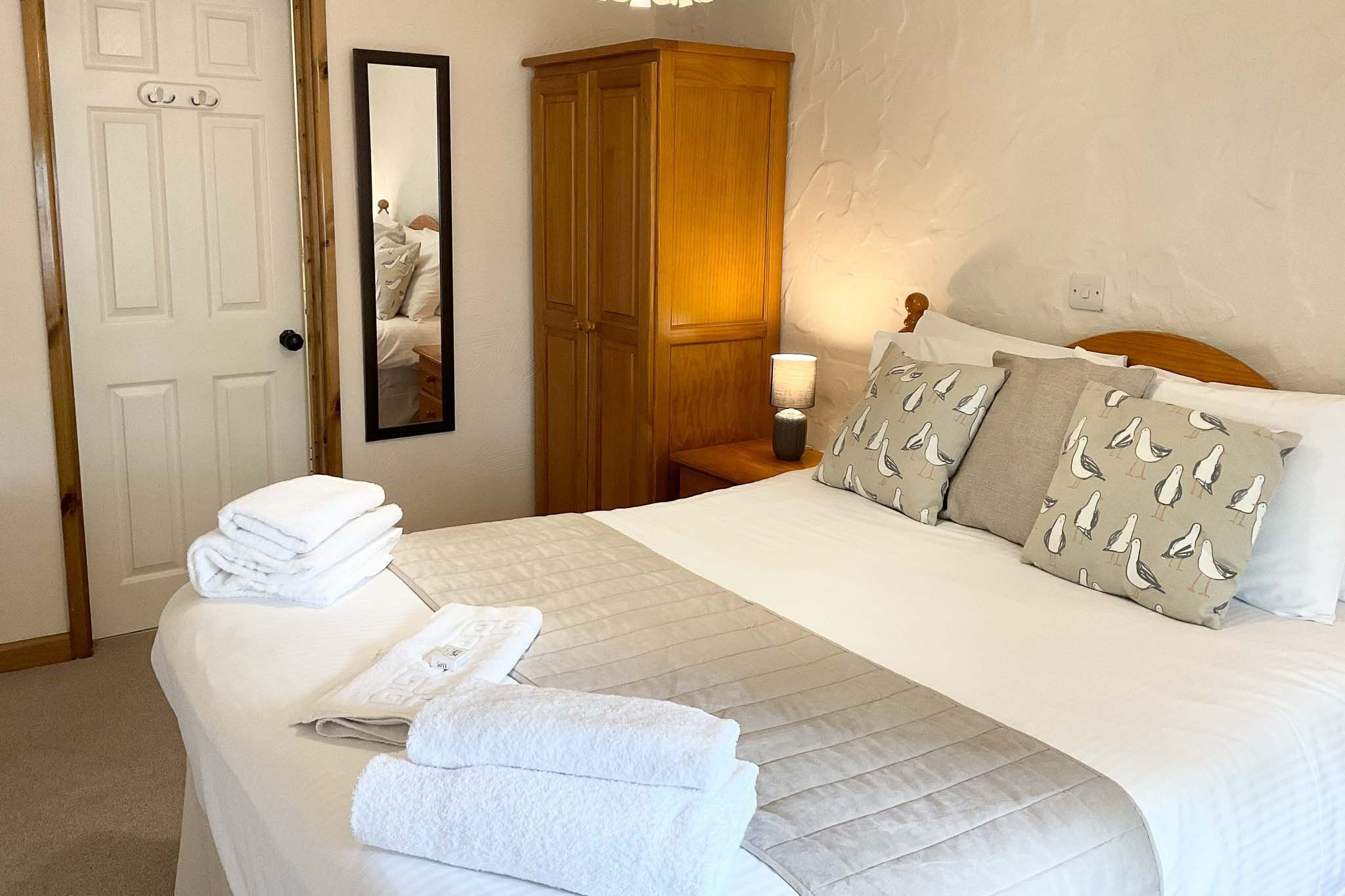 A neatly made bed with pillows and folded towels, a wooden wardrobe, and a mirror on the wall.