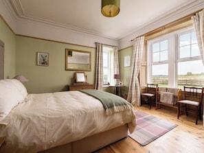Ein Schlafzimmer mit einem großen Bett, einem kleinen Tisch, einem Stuhl und Blick ins Freie durch ein Fenster.
