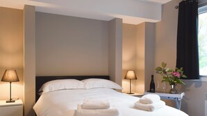 A hotel room with a bed, two bedside lamps, a nightstand, a vase with flowers, and a window with curtains.