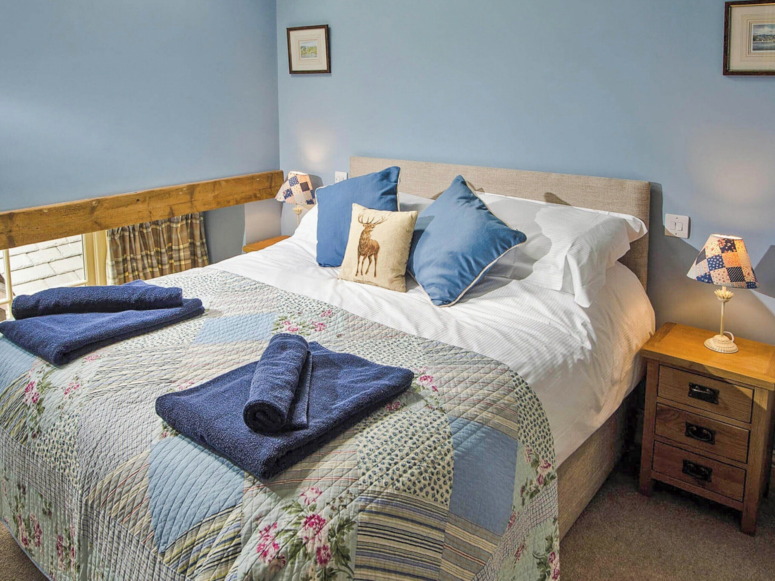 A neatly made bed with a floral quilt, blue pillows, and a deer-themed pillow. There are two rolled towels on the bed, a bedside lamp, and a wooden nightstand with drawers.