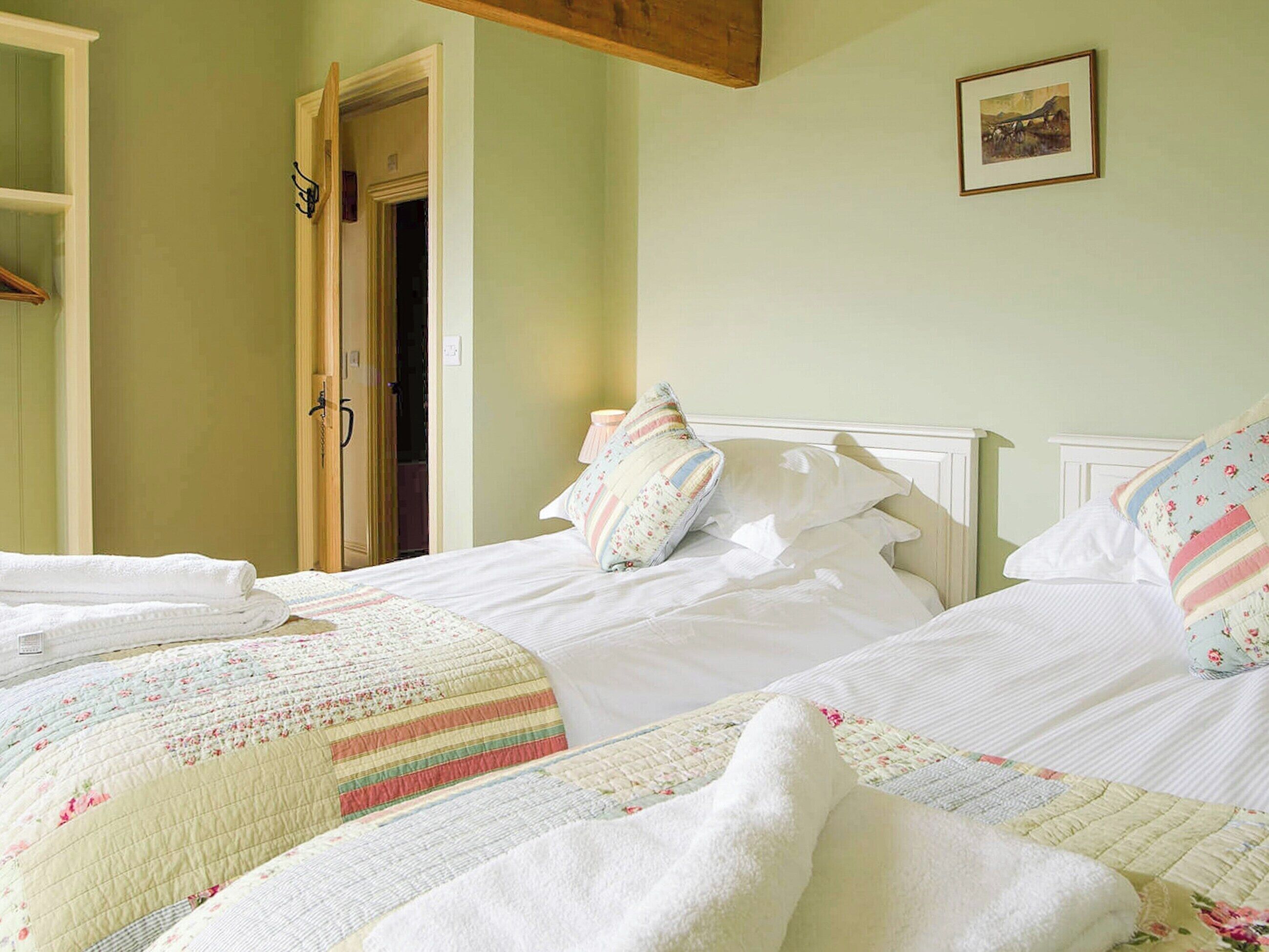 Une chambre à coucher avec deux lits, chacun recouvert d’une courtepointe à motifs floraux et doté de draps blancs. On y trouve une porte en bois et un tableau encadré accroché au mur.