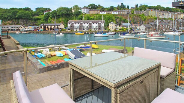 Un balcon agrémenté de meubles en osier et d’une table en verre, offrant une vue sur une marina avec des bateaux et des maisons en arrière-plan.