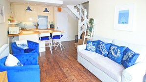 Un salon moderne avec un canapé blanc et des fauteuils bleus, une table à manger en bois avec des chaises, et un escalier menant à un niveau supérieur.