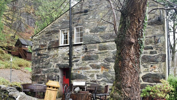 Une petite maison en pierre avec une porte rouge, un banc et un espace salon extérieur.