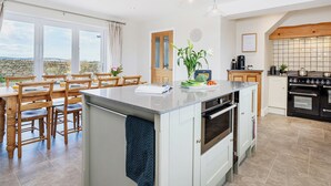 Une cuisine moderne avec un îlot central, une table à manger en bois et des chaises, une vue dégagée grâce à de grandes fenêtres et une horloge au mur.