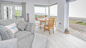A modern living room with a grey sofa, wooden dining table, and chairs. There is a large window providing a view of the outdoors. - Captains Beach House (Aberdesach)