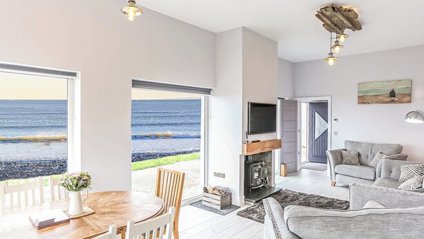 A spacious living area with a large window overlooking a beach, a cozy seating area with a fireplace, and a dining table with chairs. - Captains Beach House (Aberdesach)